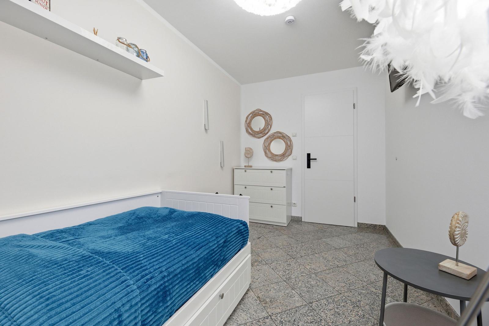 Schlafzimmer mit weißem Bett, blauer Bettdecke, Schrank und Wanddekoration.