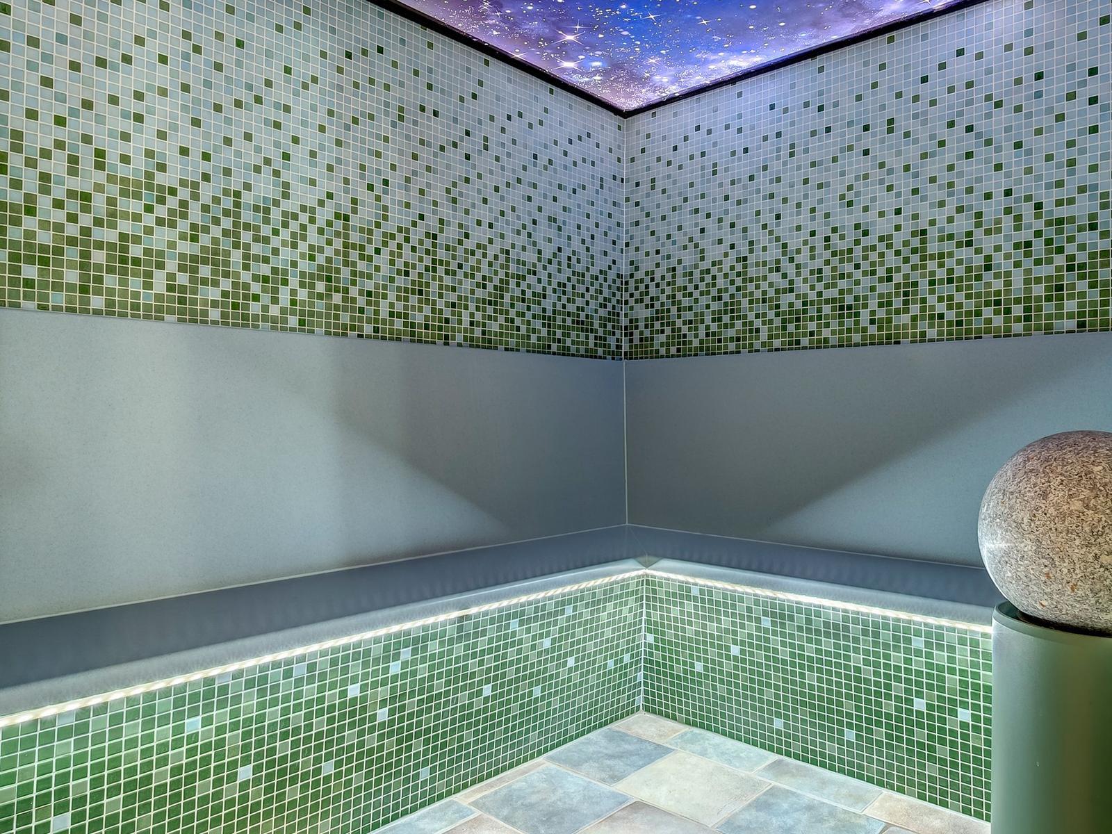 Raum mit grünen Mosaikfliesen, sternenhimmelartigem Deckenlicht und Steinball auf Sockel.