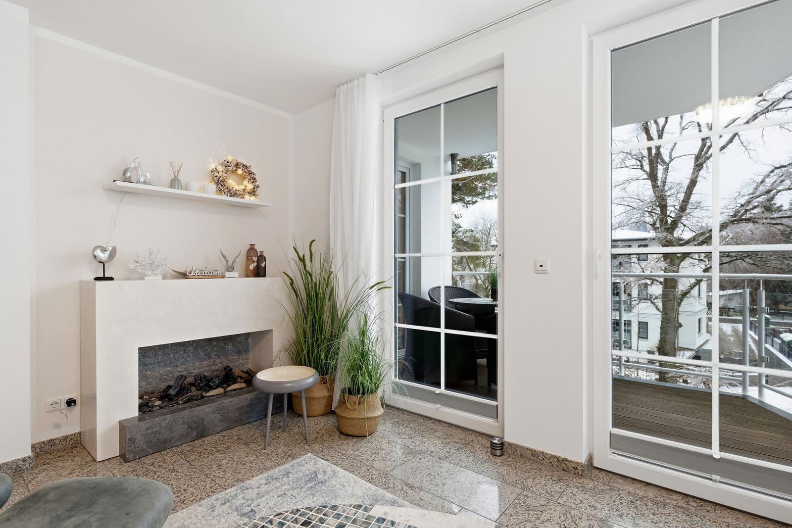 Wohnzimmer mit Kamin, Pflanzen und Terrassentür. Blick auf Balkon mit Garten.
