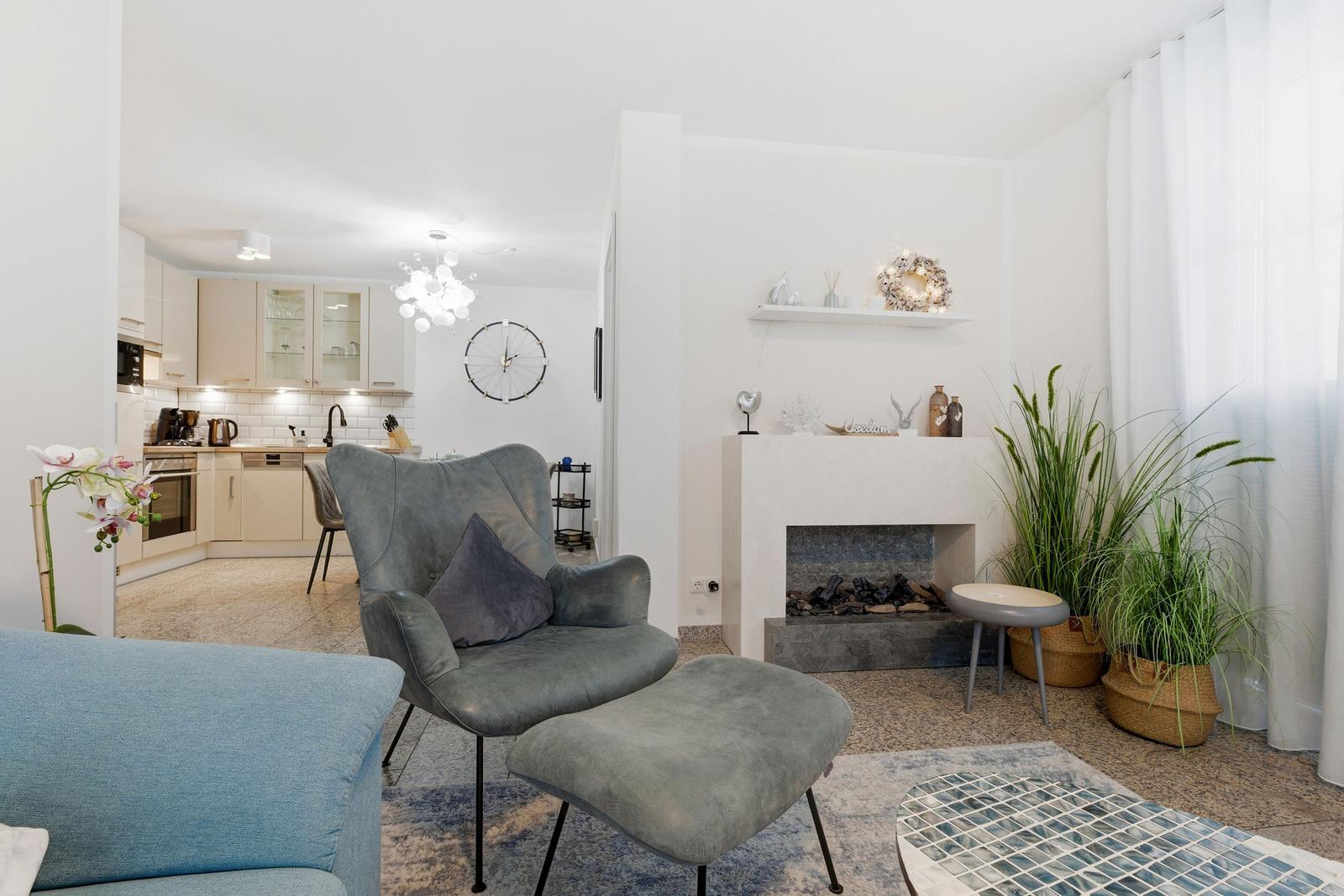 Wohnzimmer mit Sessel, Kamin und Küchenbereich im Hintergrund.