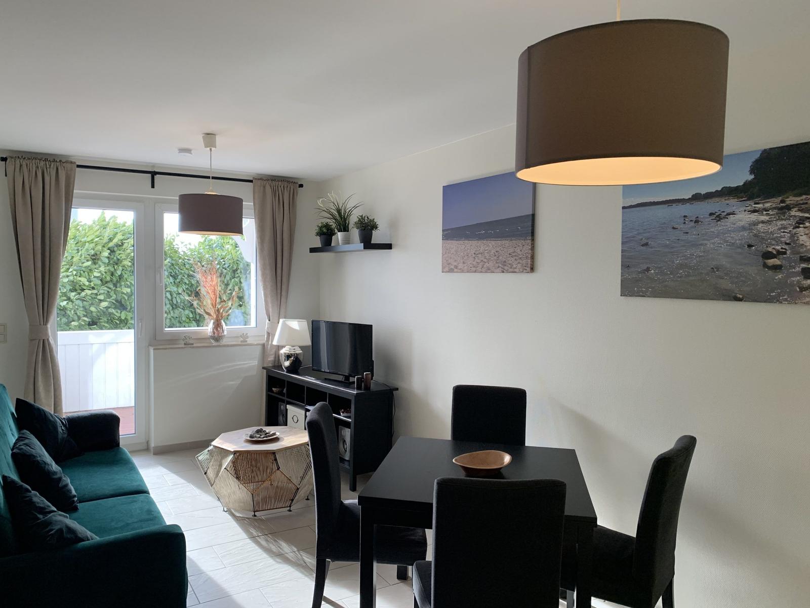 Wohn- und Essbereich mit Sofa, Tisch und Stühlen, Fenster mit Blick auf Grünflächen.