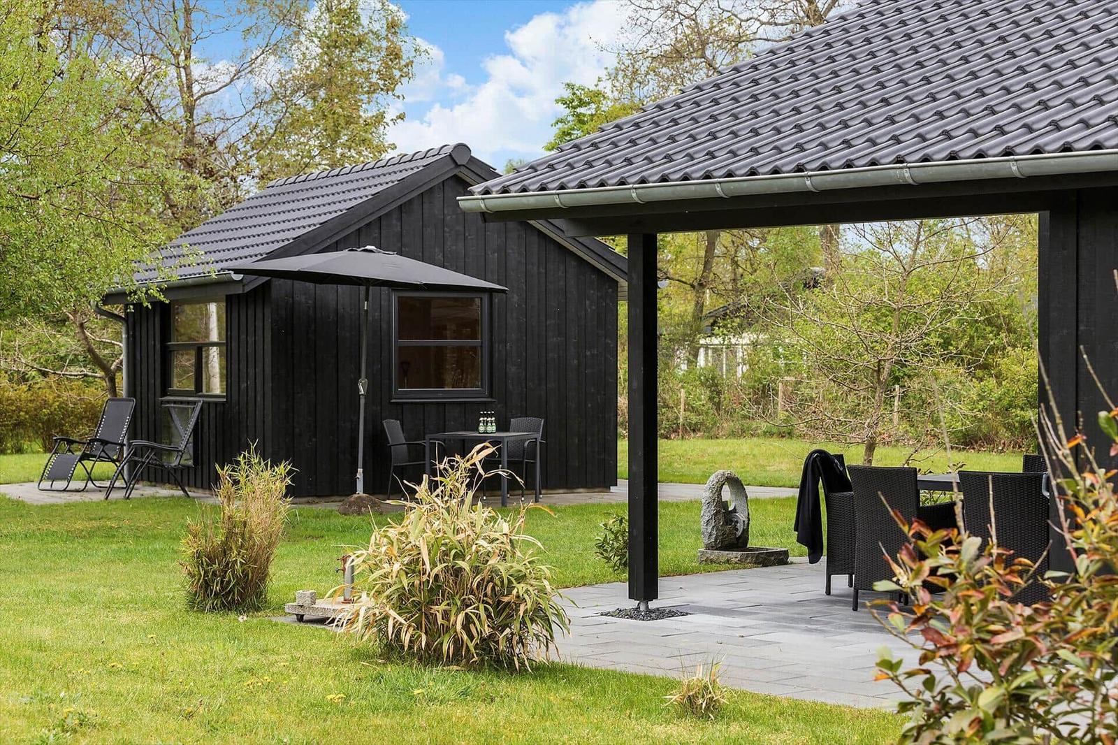 Schwarze Holzhütte mit Terrasse und Gartenmöbeln unter einem Dach.