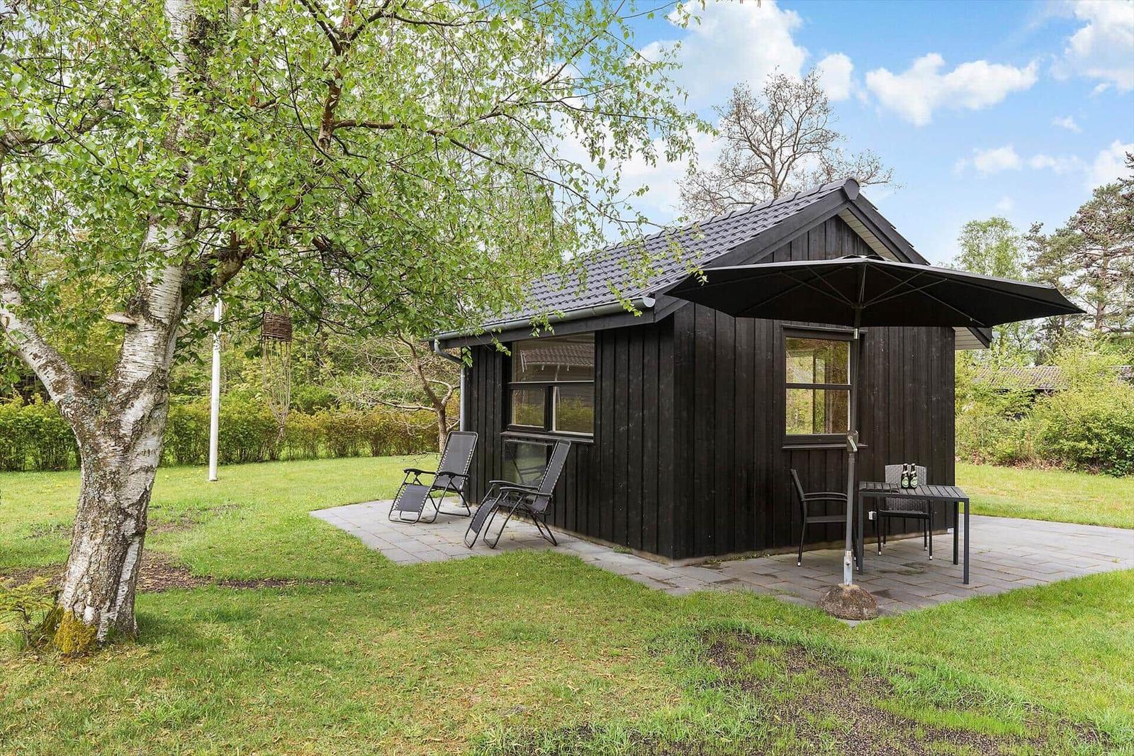 Schwarze Holzhütte mit Terrasse, Sitzgelegenheiten und Sonnenschutz im Grünen.