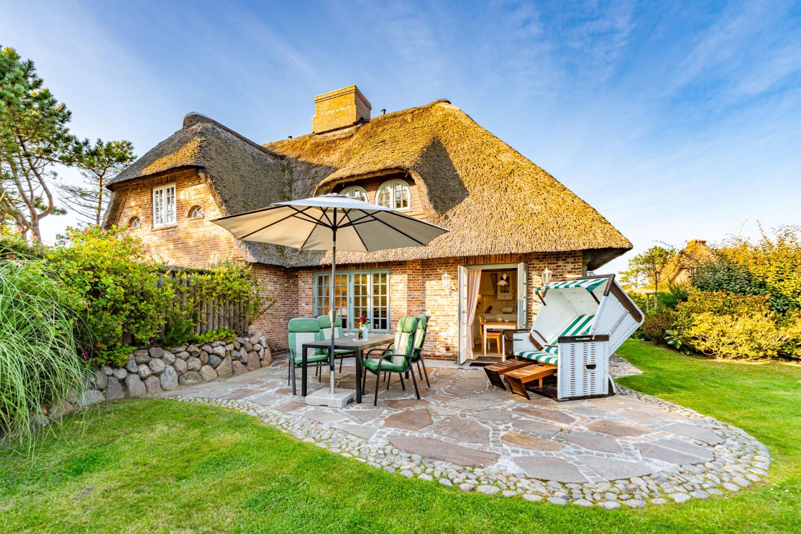 Haus mit Strohdach, Terrasse mit Tisch, Stühlen und Strandkorb.