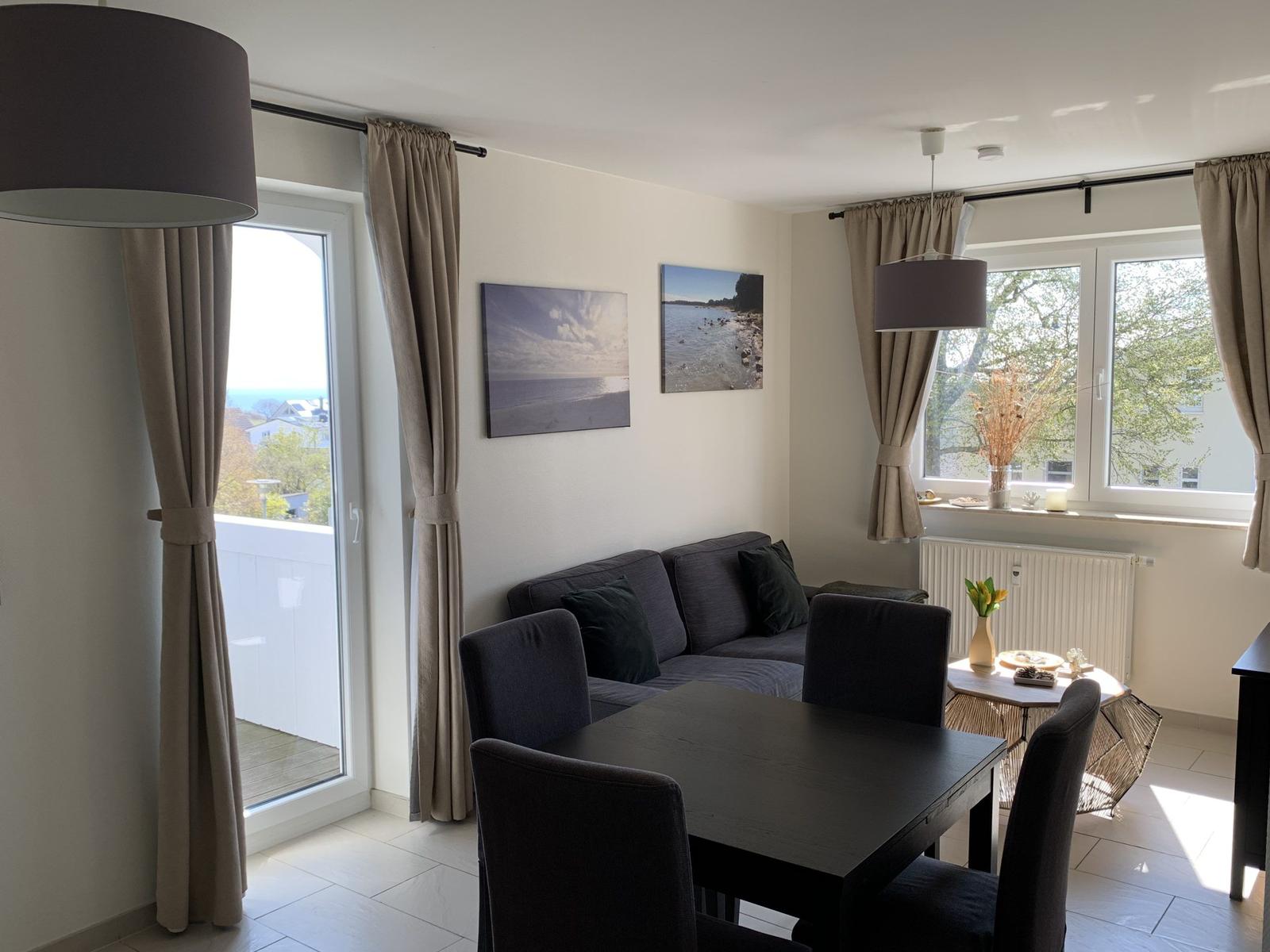 Wohn- und Essbereich mit Sofa, Tisch und Stühlen. Fenster und Balkontür mit Blick.