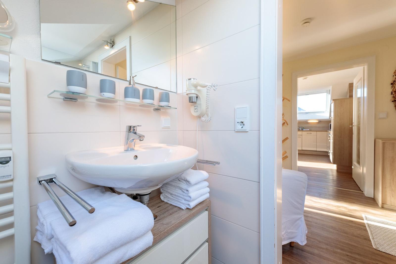 Badezimmer mit Waschbecken, Spiegel und Handtuchhalter. Blick in die Küche.