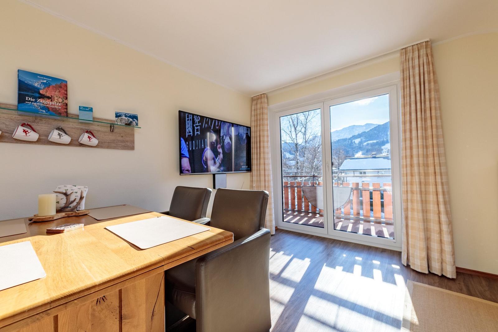 Ein Raum mit Tisch, Stühlen, Fernseher und Balkonblick auf Berge.