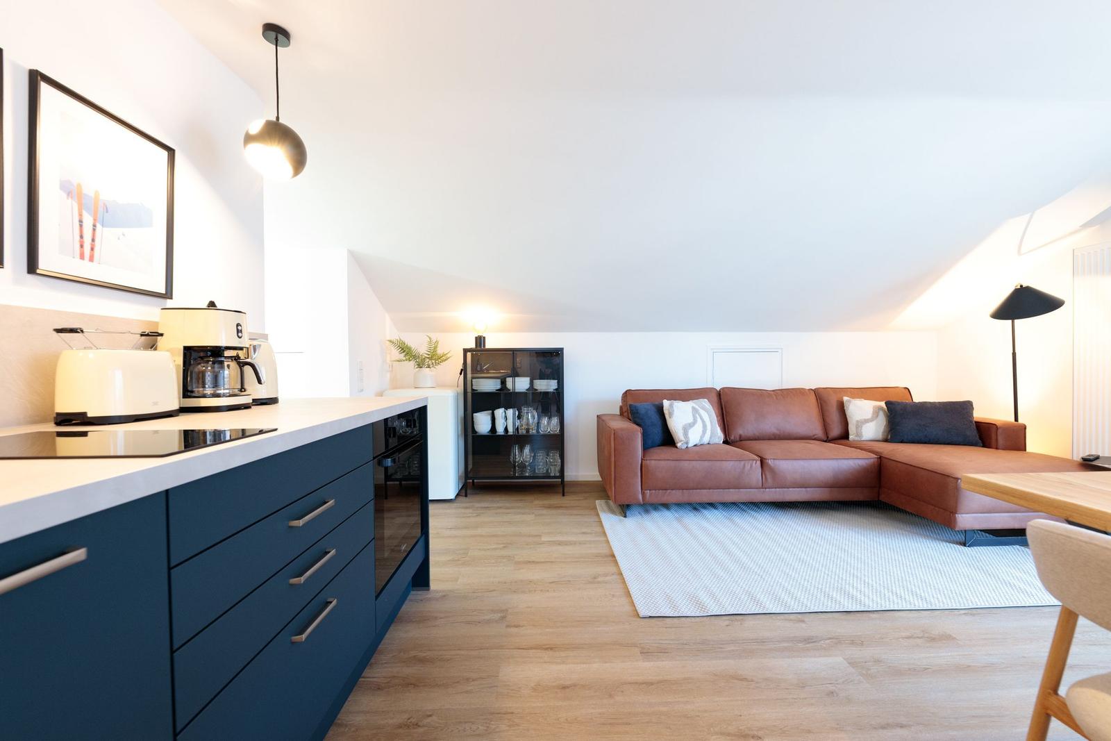 Wohn- und Küchenbereich mit braunem Sofa und dunkelblauen Schränken.