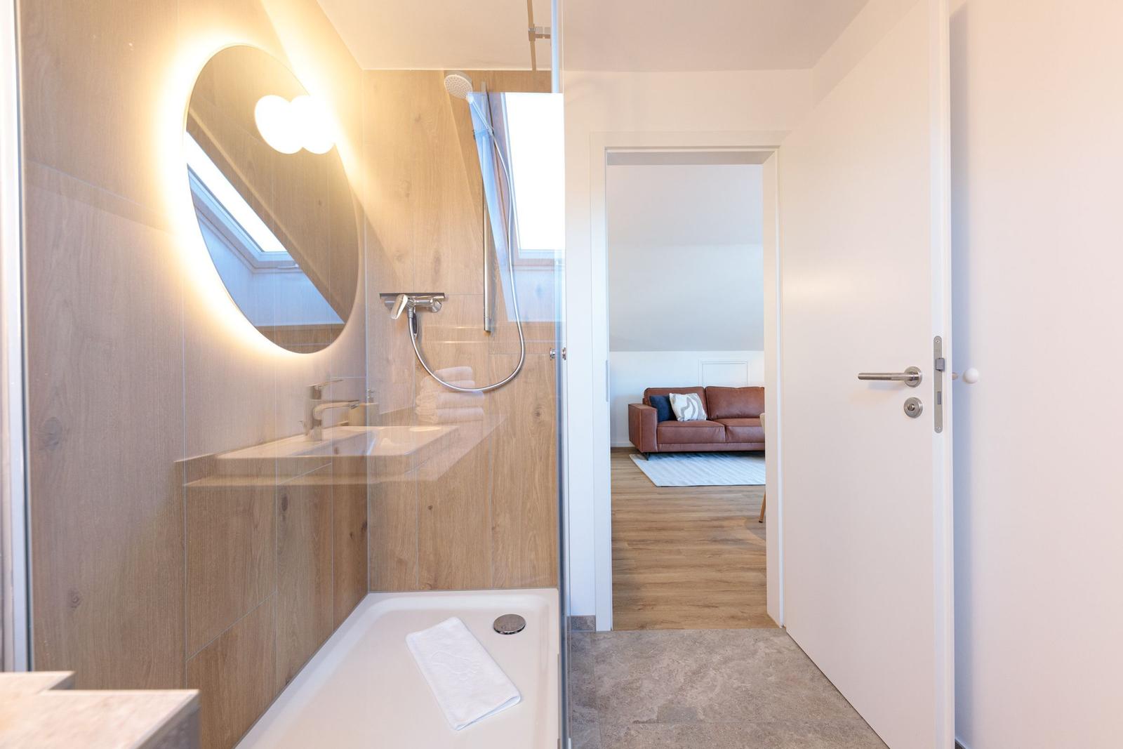 Badezimmer mit Dusche und Spiegel. Durchgang zum Wohnzimmer mit Sofa.