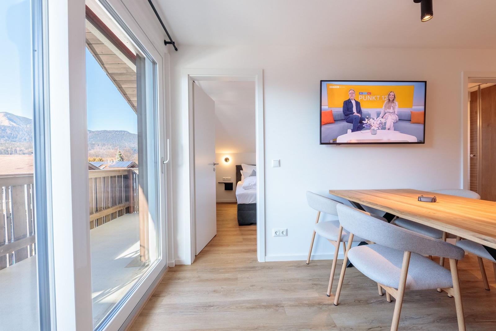Wohnbereich mit Essplatz, TV und Blick auf Balkon mit Berglandschaft.