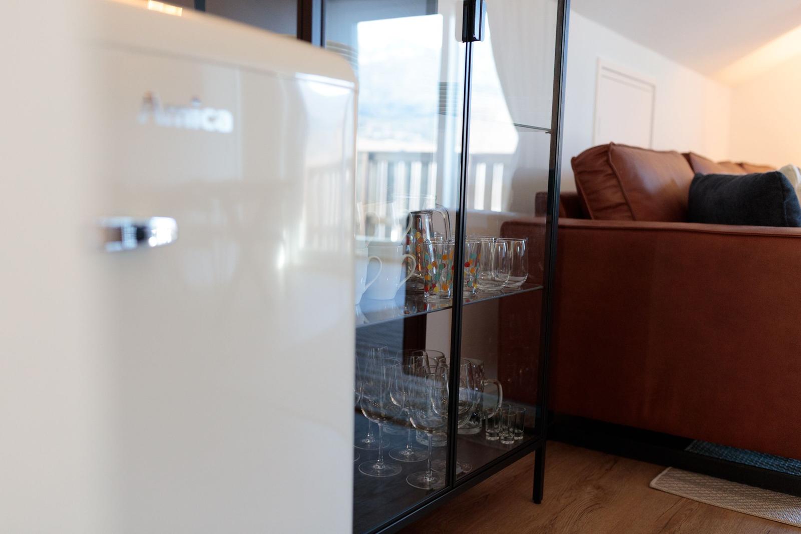 Weißer Kühlschrank neben Glasregal mit Gläsern und Sofa im Hintergrund.