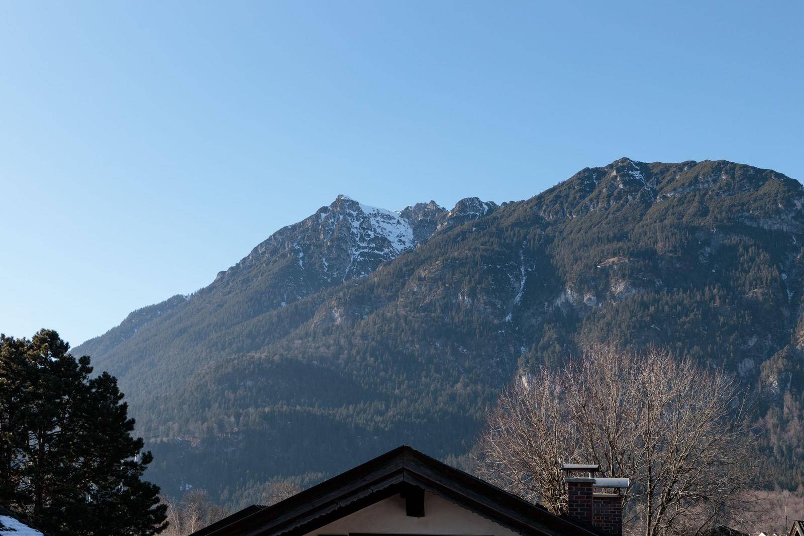 Schneebedeckte Berge mit Wald und Hausdach im Vordergrund.