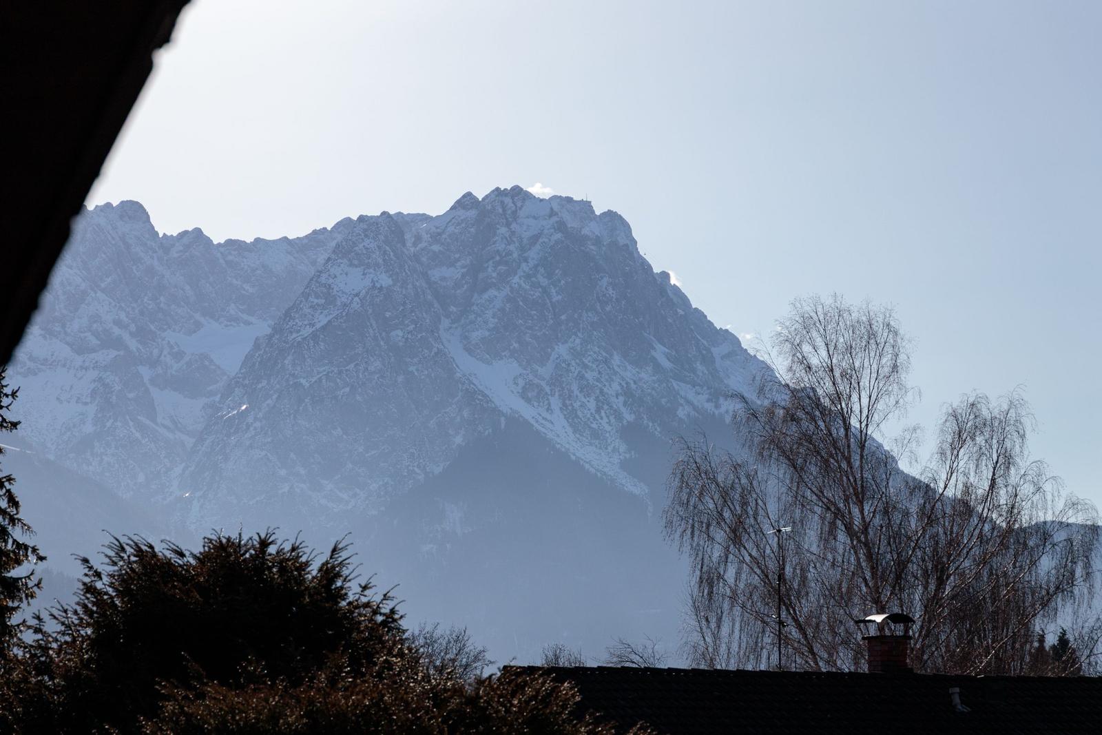 Schneebedeckte Berge sind vom Fenster aus sichtbar, umgeben von Bäumen und einem Dach.