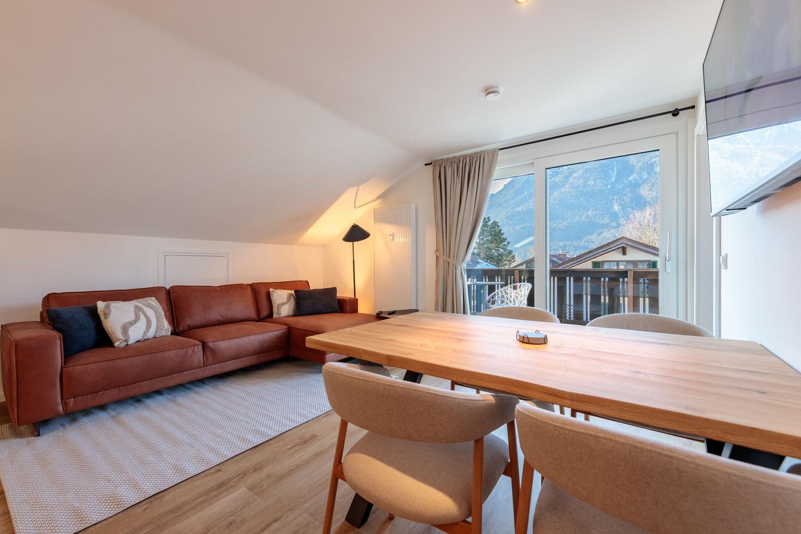 Wohn- und Essbereich mit Sofa, Tisch und Blick auf Balkon und Berge.