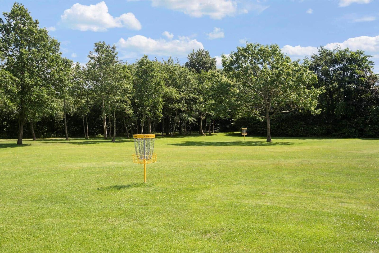 Yellow disc golf basket on green grass before trees under blue sky.