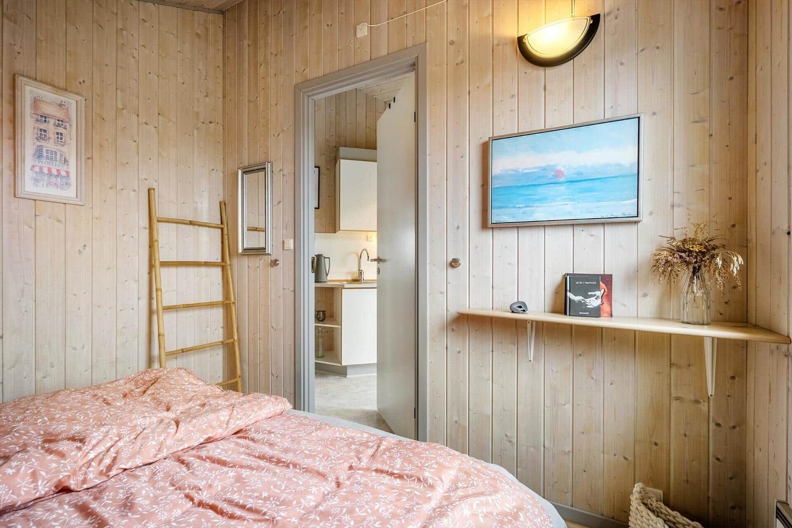 Bedroom with wooden walls, bed, and TV. View into the kitchen through doorway.