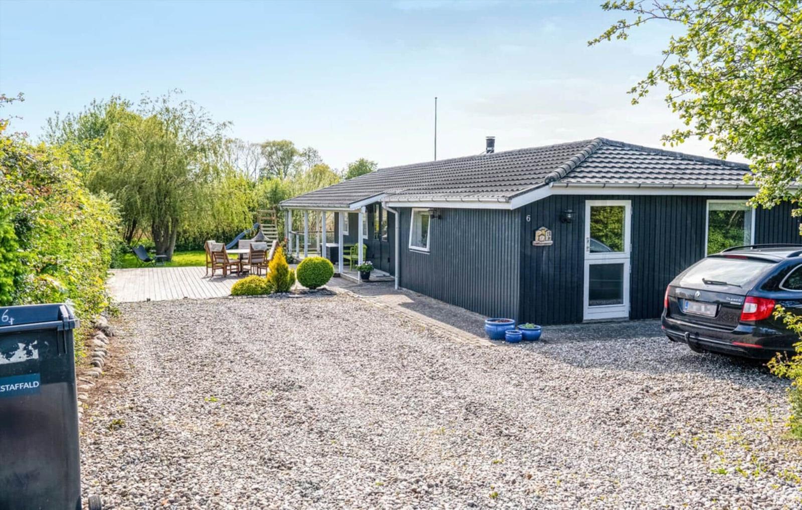 A house with dark siding, patio, and garden. A car is parked on the gravel drive.