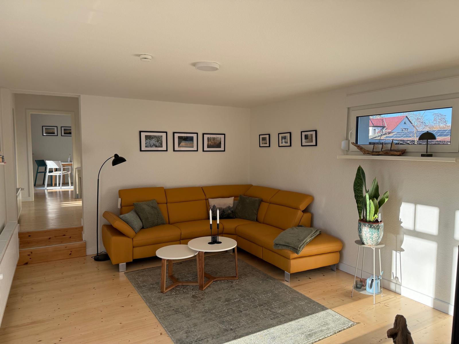 Wohnzimmer mit gelbem Sofa, Tischen und Pflanze. Durchgang zur Küche.