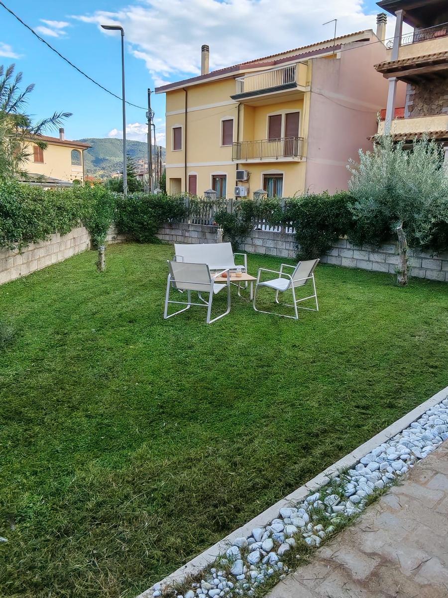 Green garden with seating area and stone wall. Background: yellow house with balcony.