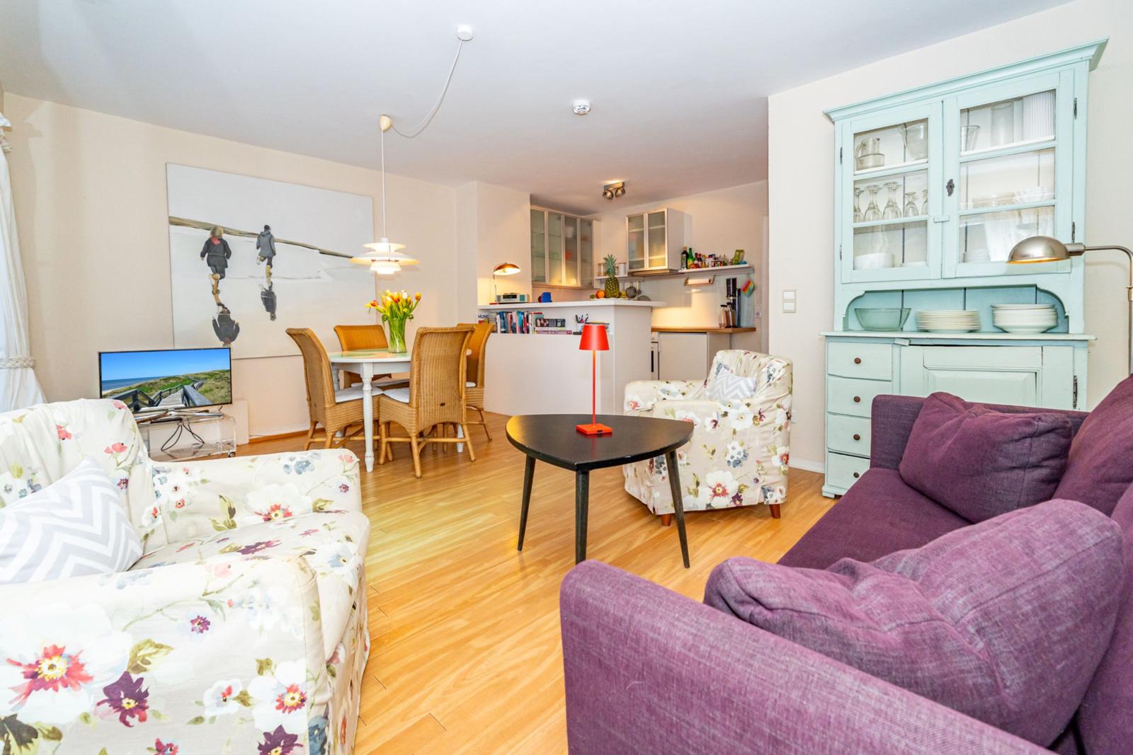 Wohnzimmer mit Sofa, Sessel und Esstisch. Küche im Hintergrund. Holzboden und Küchenschränke.