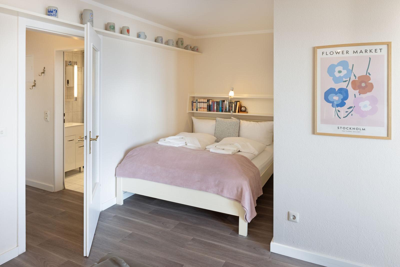 Schlafzimmer mit Bett, Wandregal und Bild. Tür führt in Bad.