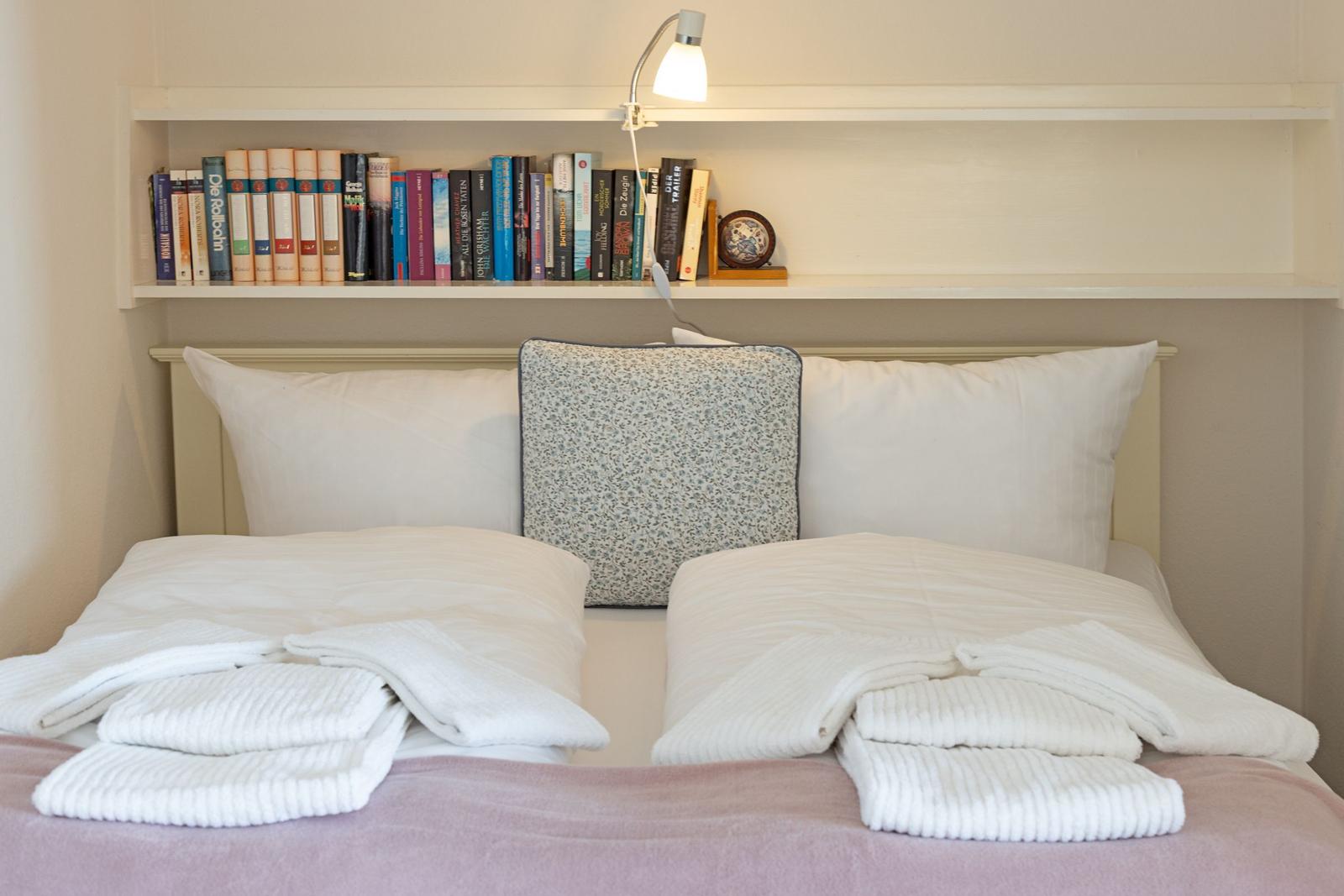 Doppelbett mit weißen Kissen und gefalteten Handtüchern. Bücherregal über dem Bett.
