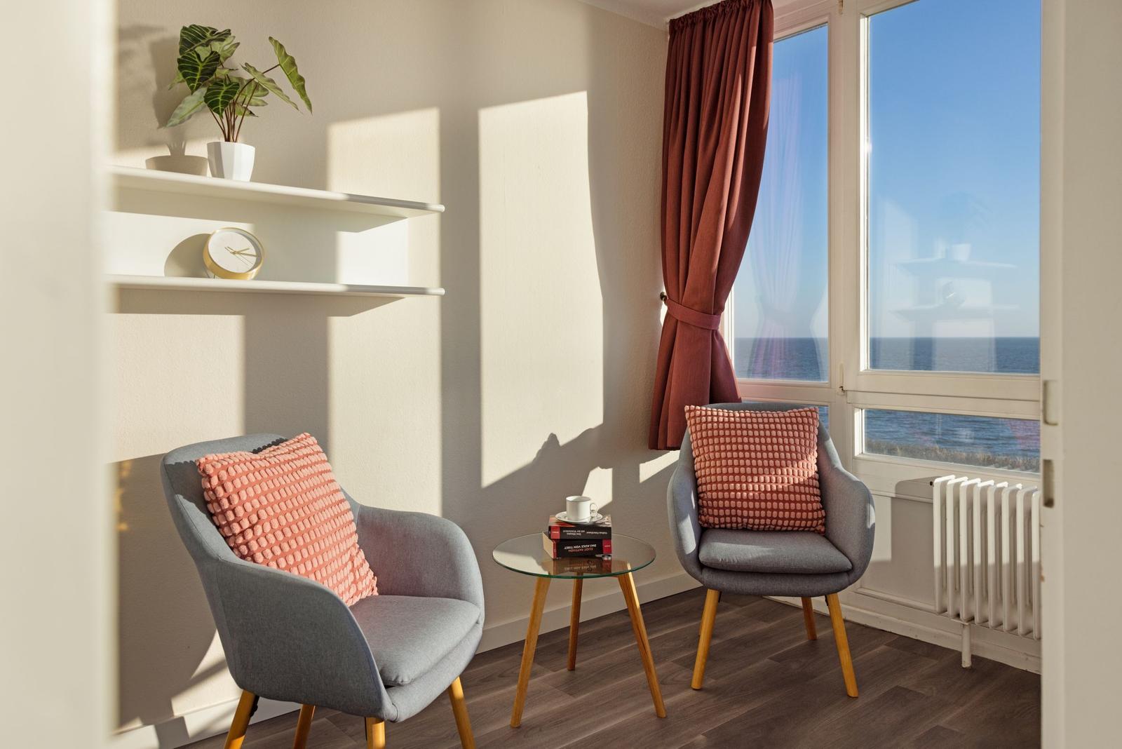 Zwei graue Sessel mit rosa Kissen stehen vor einem Fenster mit Meerblick.