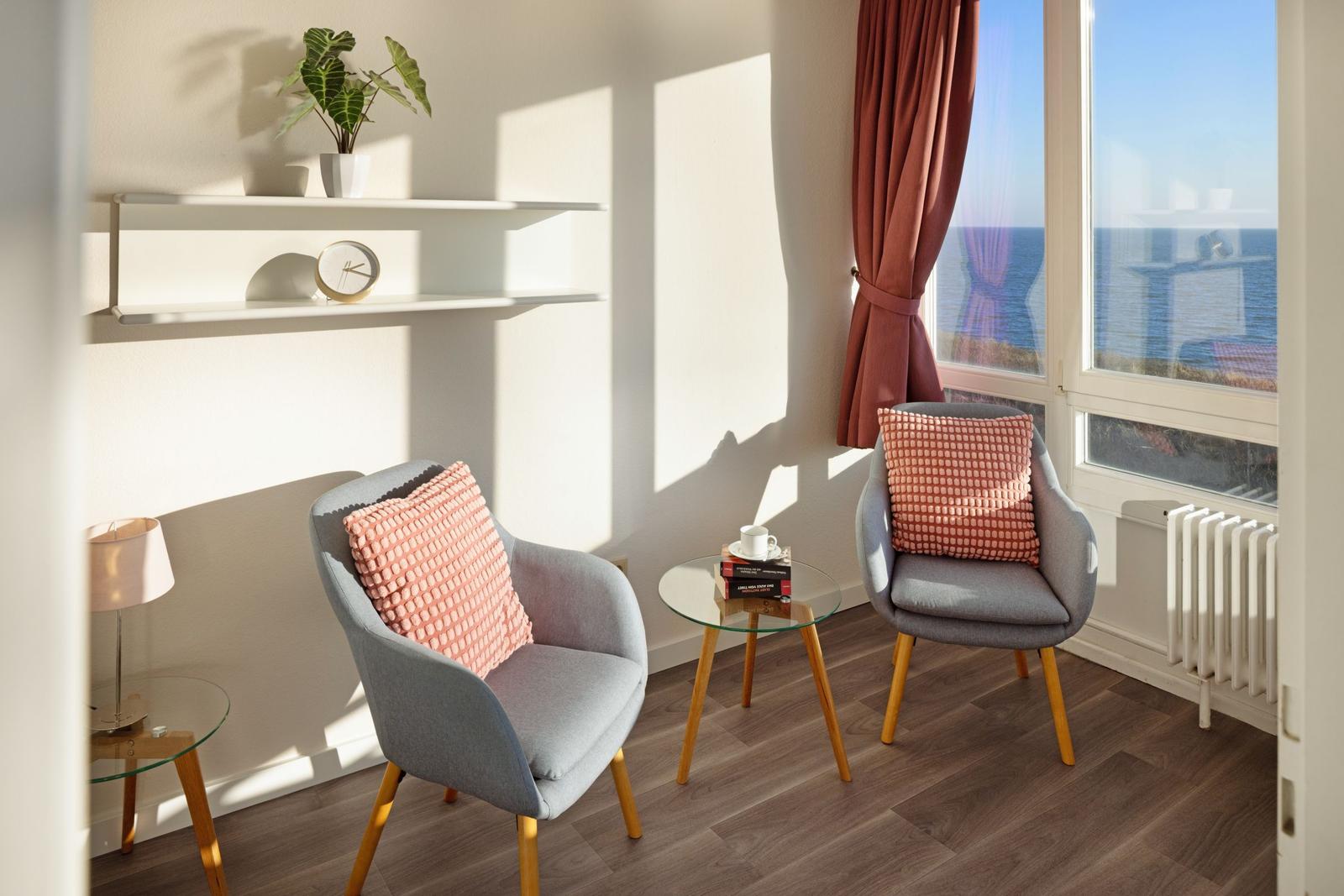 Zwei graue Sessel mit rosa Kissen stehen vor großem Fenster mit Meerblick.