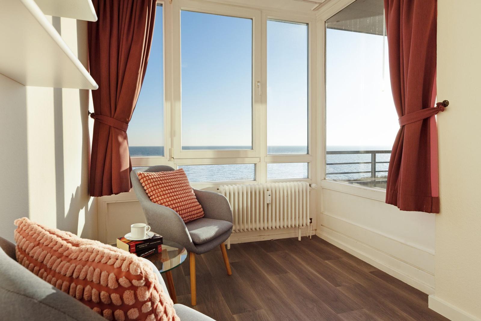 Ein Raum mit Blick auf das Meer, graue Sitzmöbel, rote Vorhänge und Holzboden.