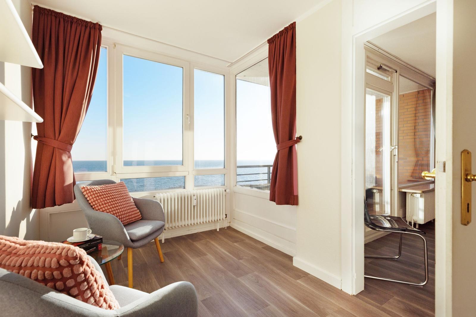 Wohnzimmer mit Meerblick, graue Sessel, rote Vorhänge und Holzboden.