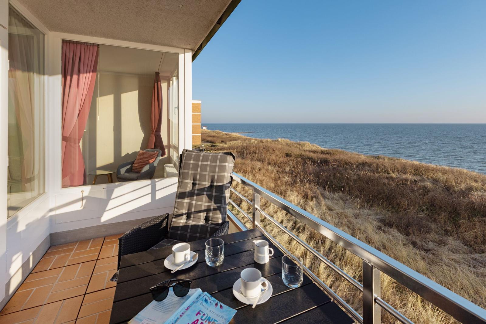 Terrasse mit Tisch, Stühlen und Meerblick. Im Hintergrund ist ein Fenster mit Vorhängen zu sehen.