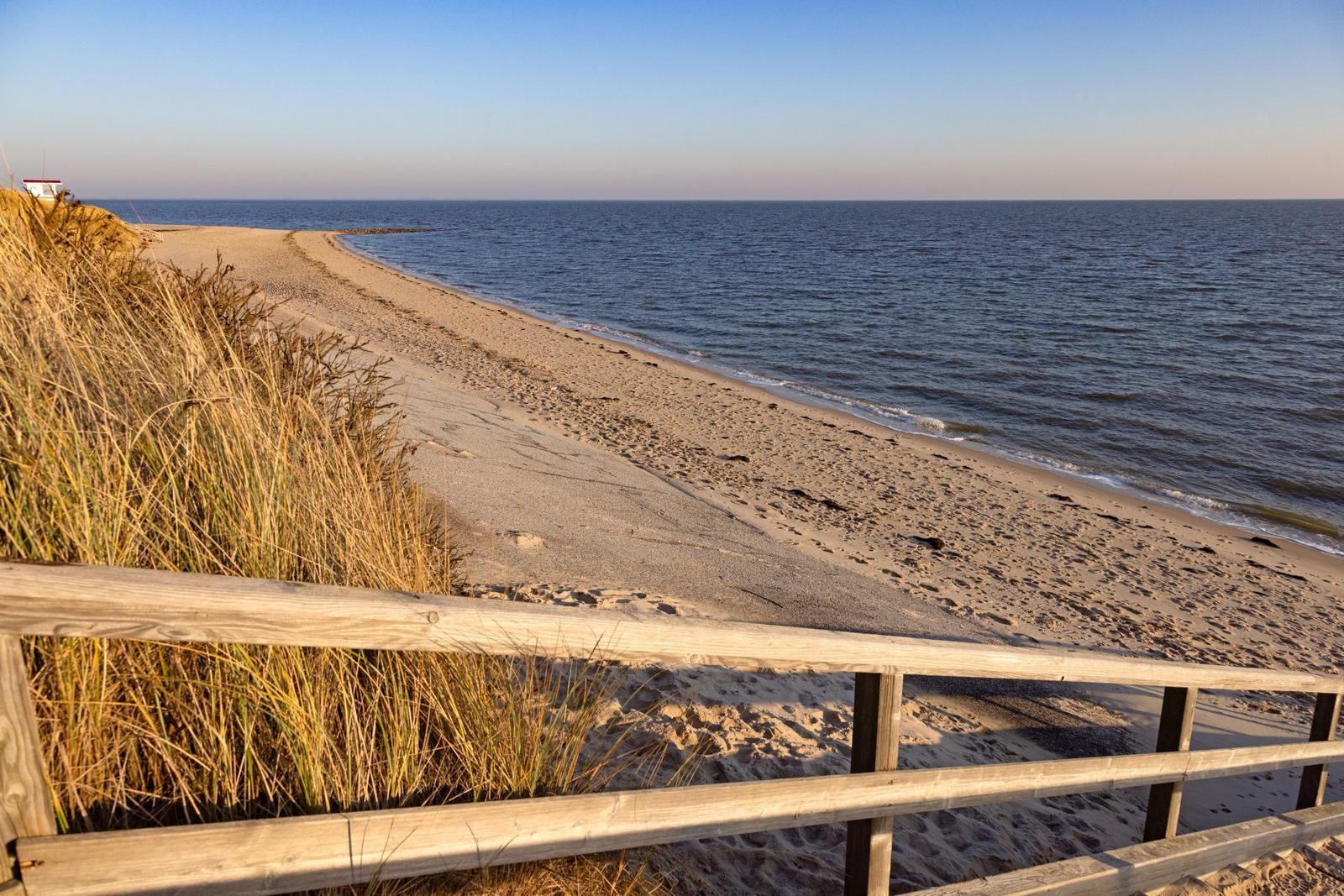 Holzgeländer führt zum Strand. Sandstrand mit Wellen und Gras.
