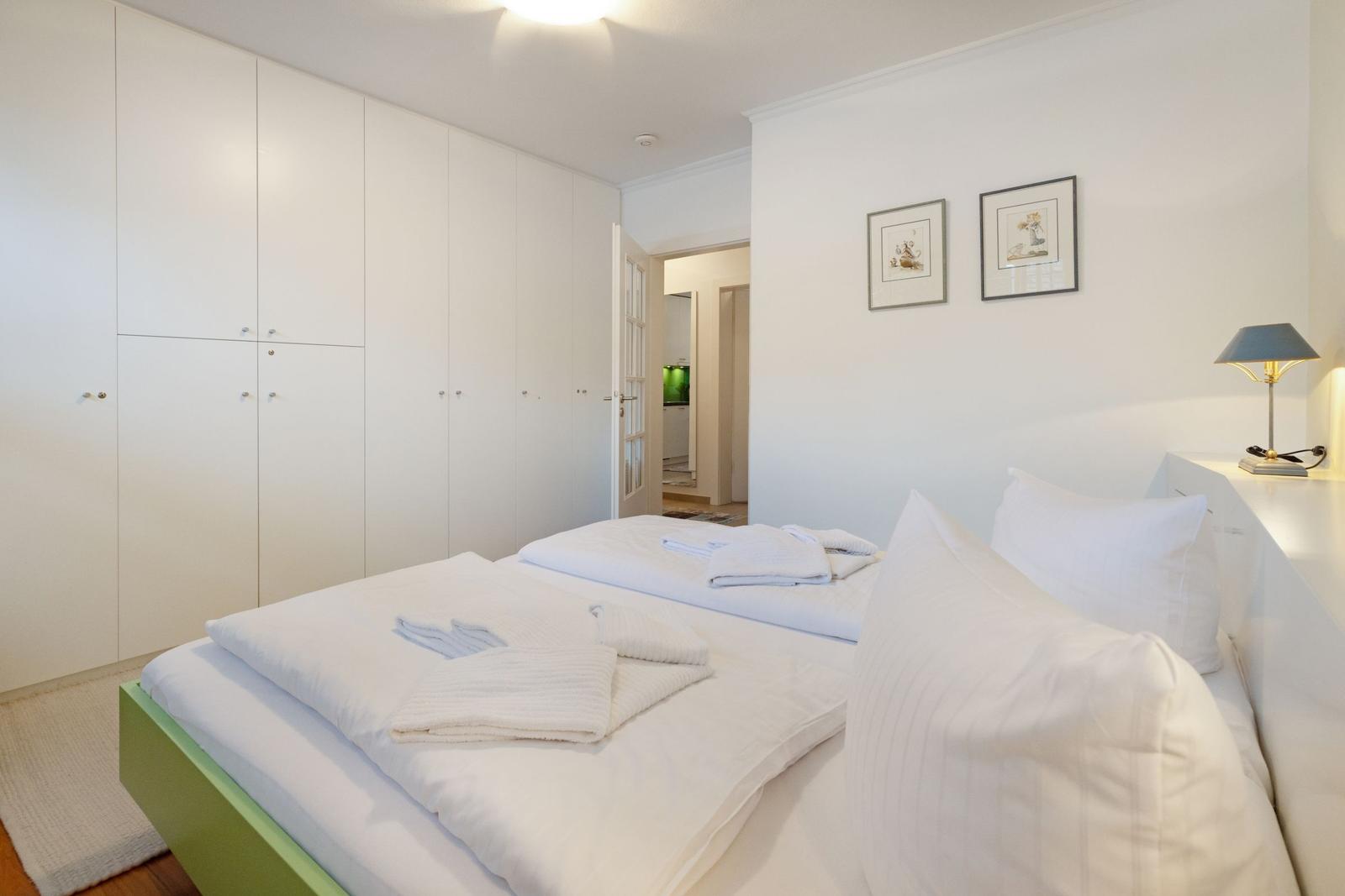 Schlafzimmer mit weißem Bett, Kleiderschrank und Nachttisch mit Lampe.