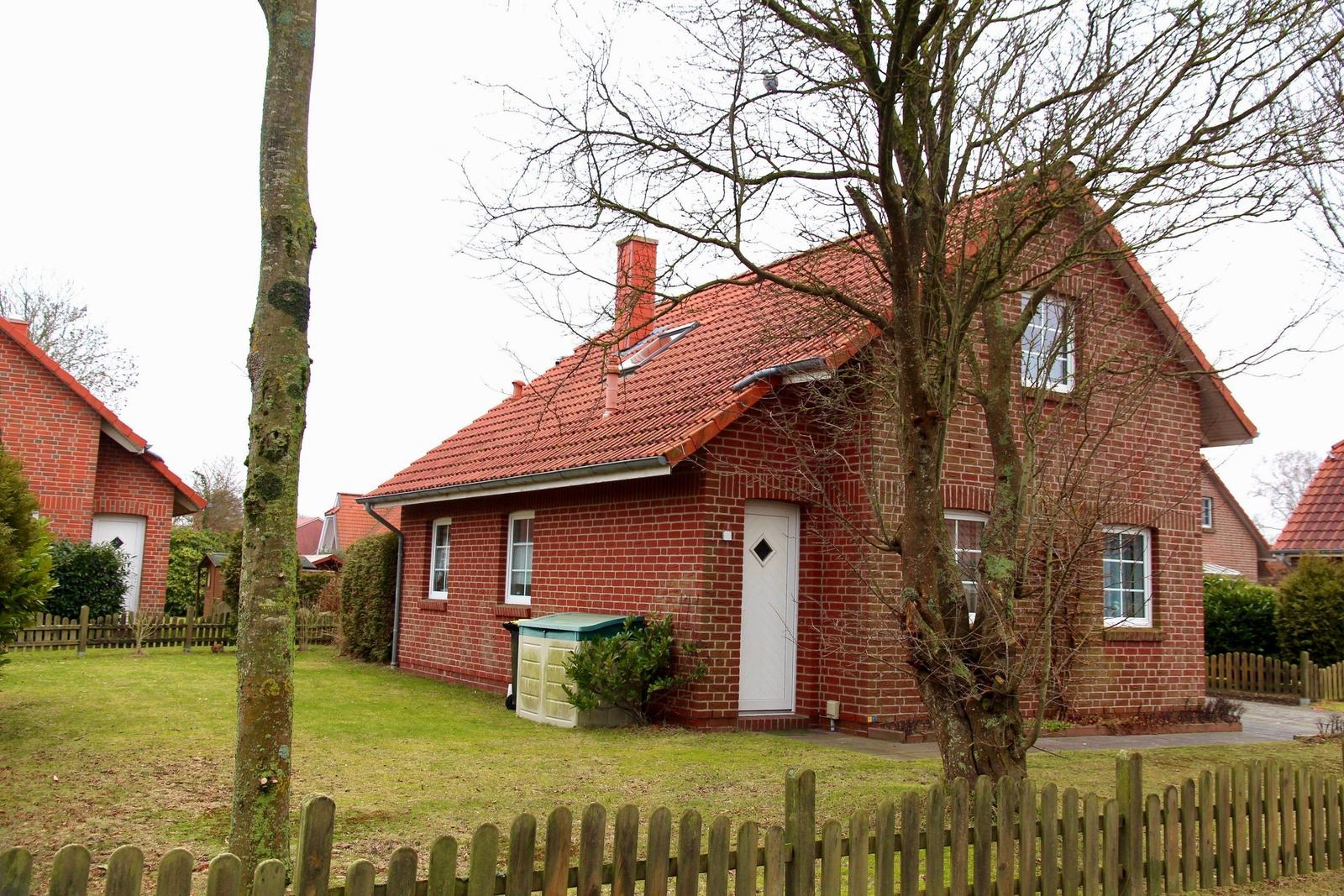 Rotes Ziegelhaus mit weißen Fenstern und Tür. Vorgarten mit Holzzaun und Bäumen.