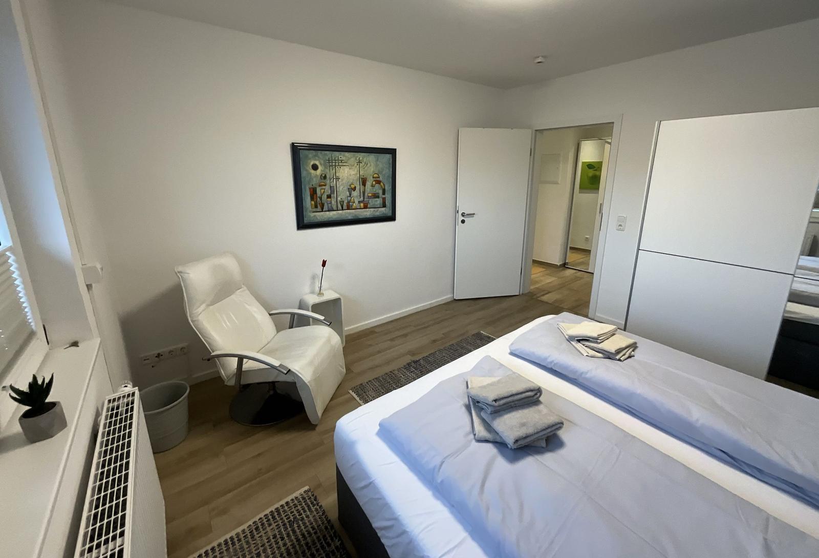 Schlafzimmer mit Bett, weißer Sessel und Wandbild.