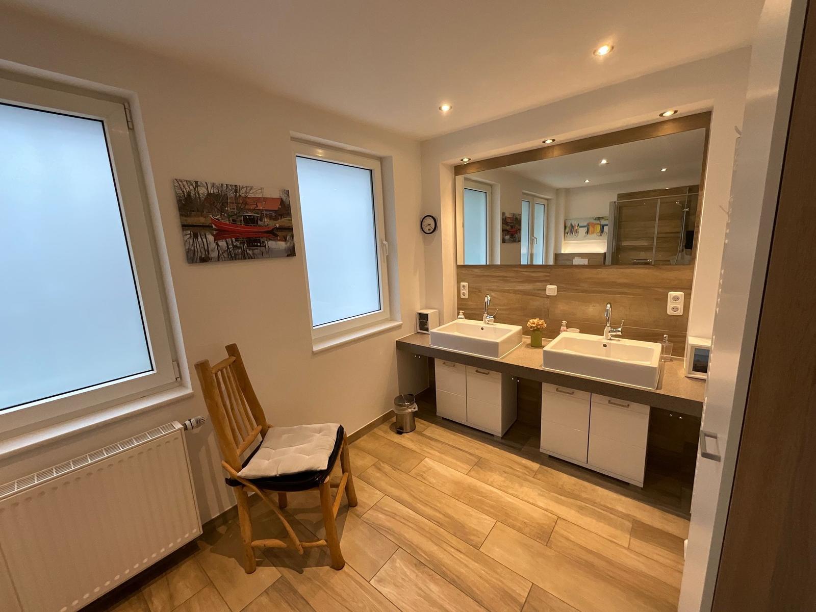 Badezimmer mit zwei Waschbecken, Spiegeln und Holzboden. Holzstuhl und Fenster sichtbar.
