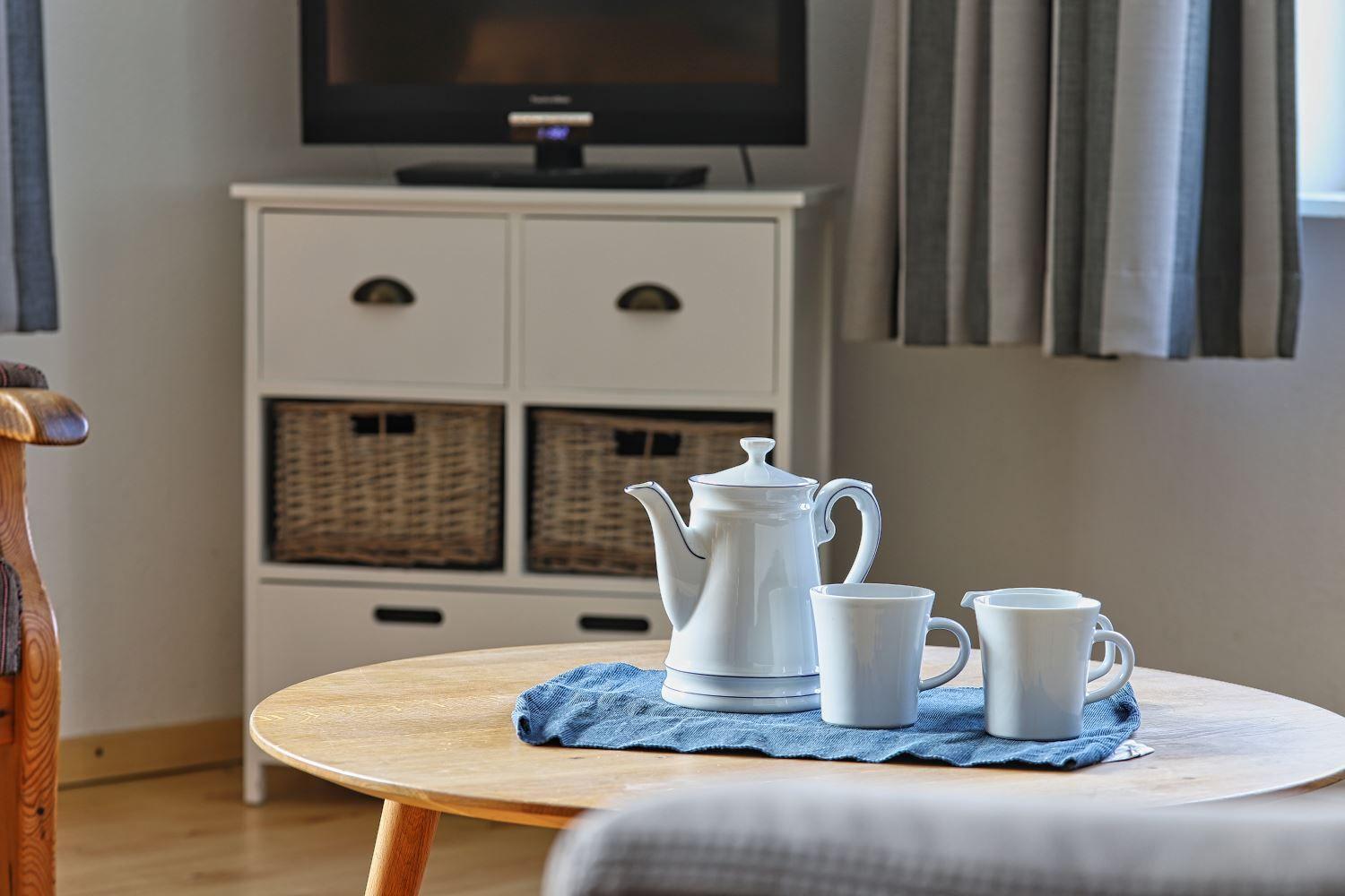 Tisch mit Teekanne und Tassen, Fernseher und Schrank im Hintergrund.