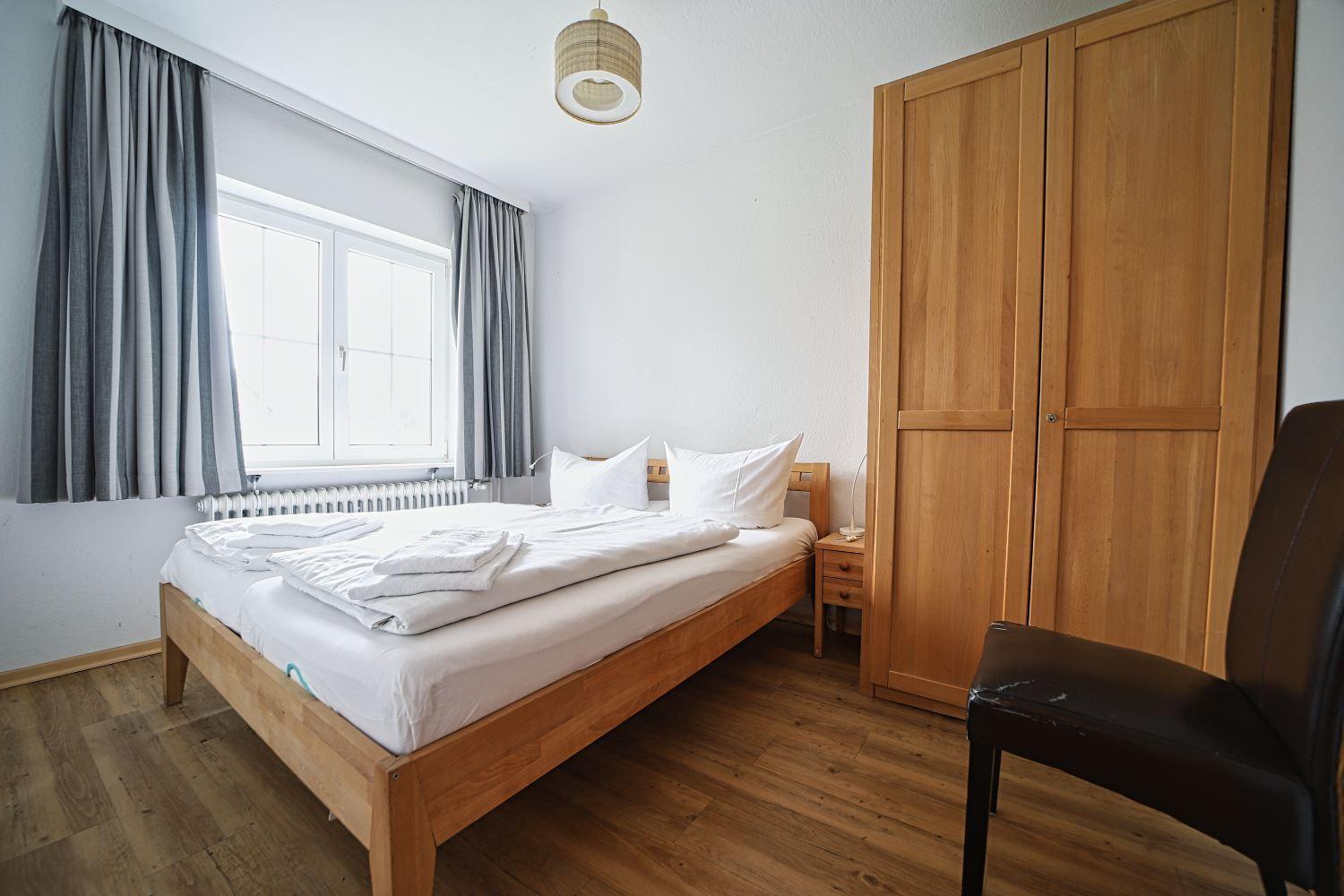 Schlafzimmer mit Holzbett, Kleiderschrank und Fenster mit Vorhängen.