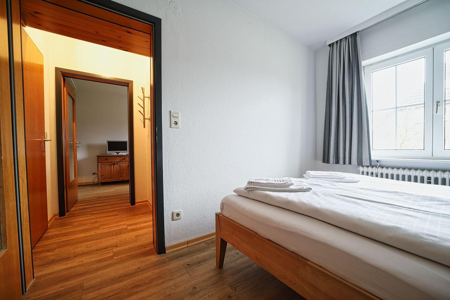 Schlafzimmer mit Bett, Fenster und Holzboden. Durchgang zu Wohnbereich mit TV.