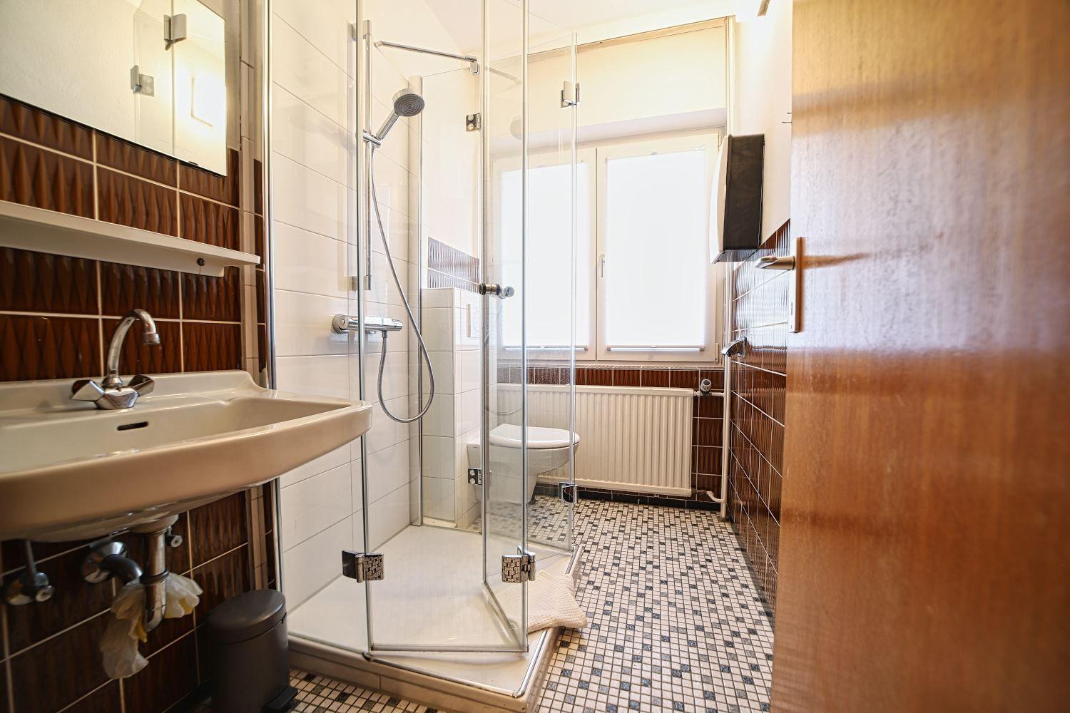 Badezimmer mit Dusche, Waschbecken und Toilette. Fliesenboden und Fenster.