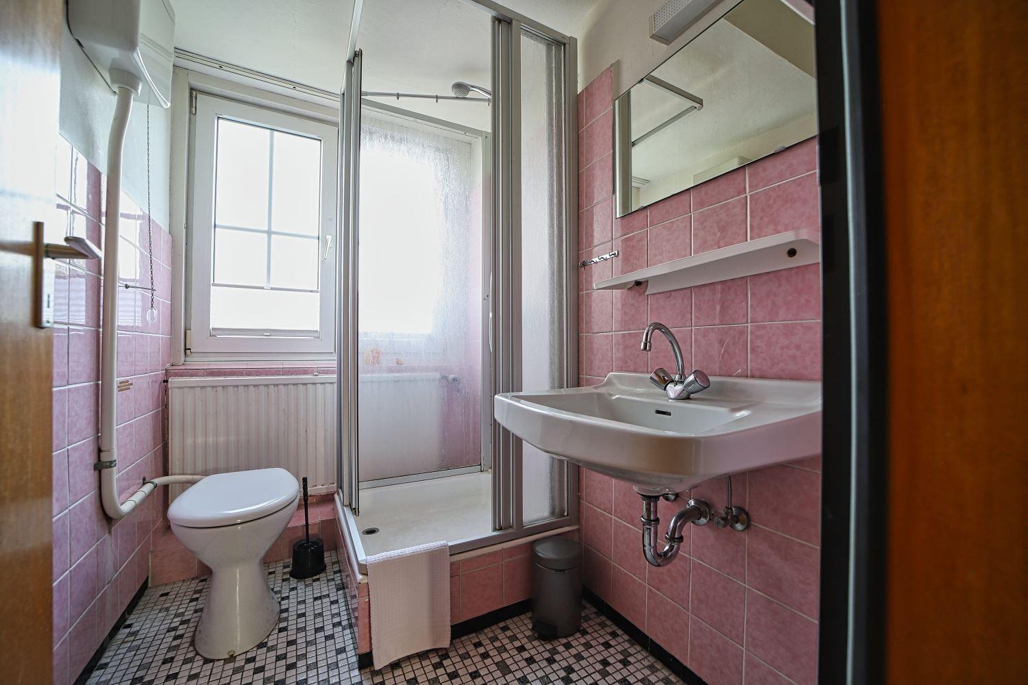 Badezimmer mit Dusche, Waschbecken und Toilette. Wandfliesen rosa, Bodenfliesen schwarz-weiß.