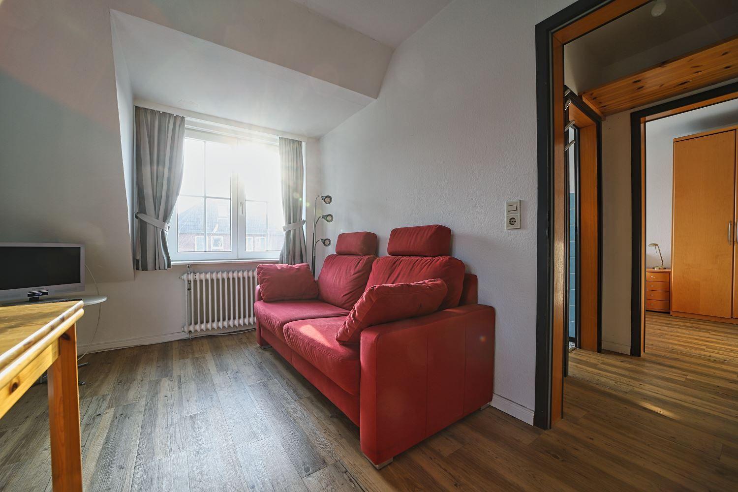 Ein Wohnzimmer mit rotem Sofa, Holzboden und Fenster mit Vorhängen.