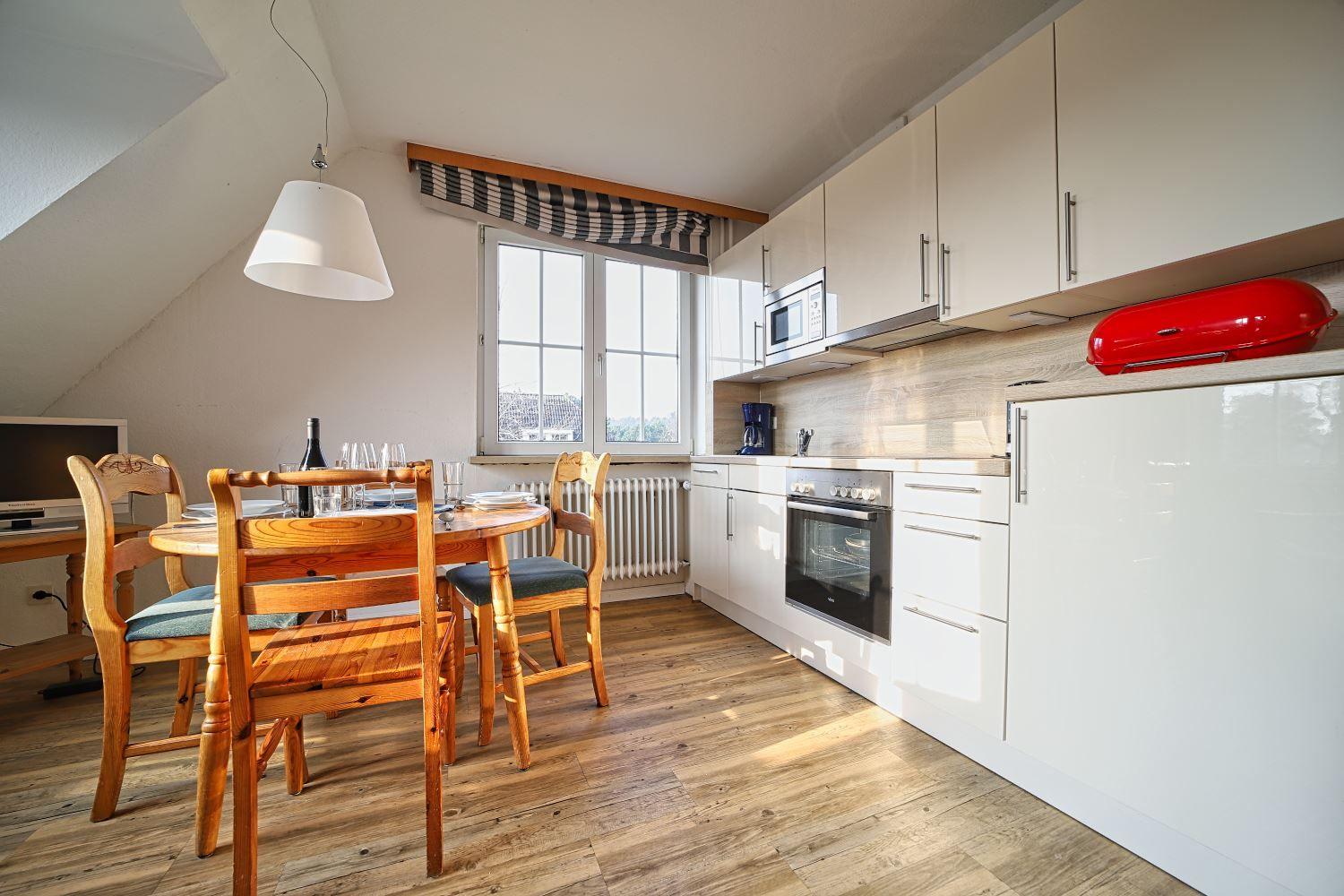 Küche mit Esstisch, Fenster und Holzboden. Weiße Schränke und rote Küchenutensilien.
