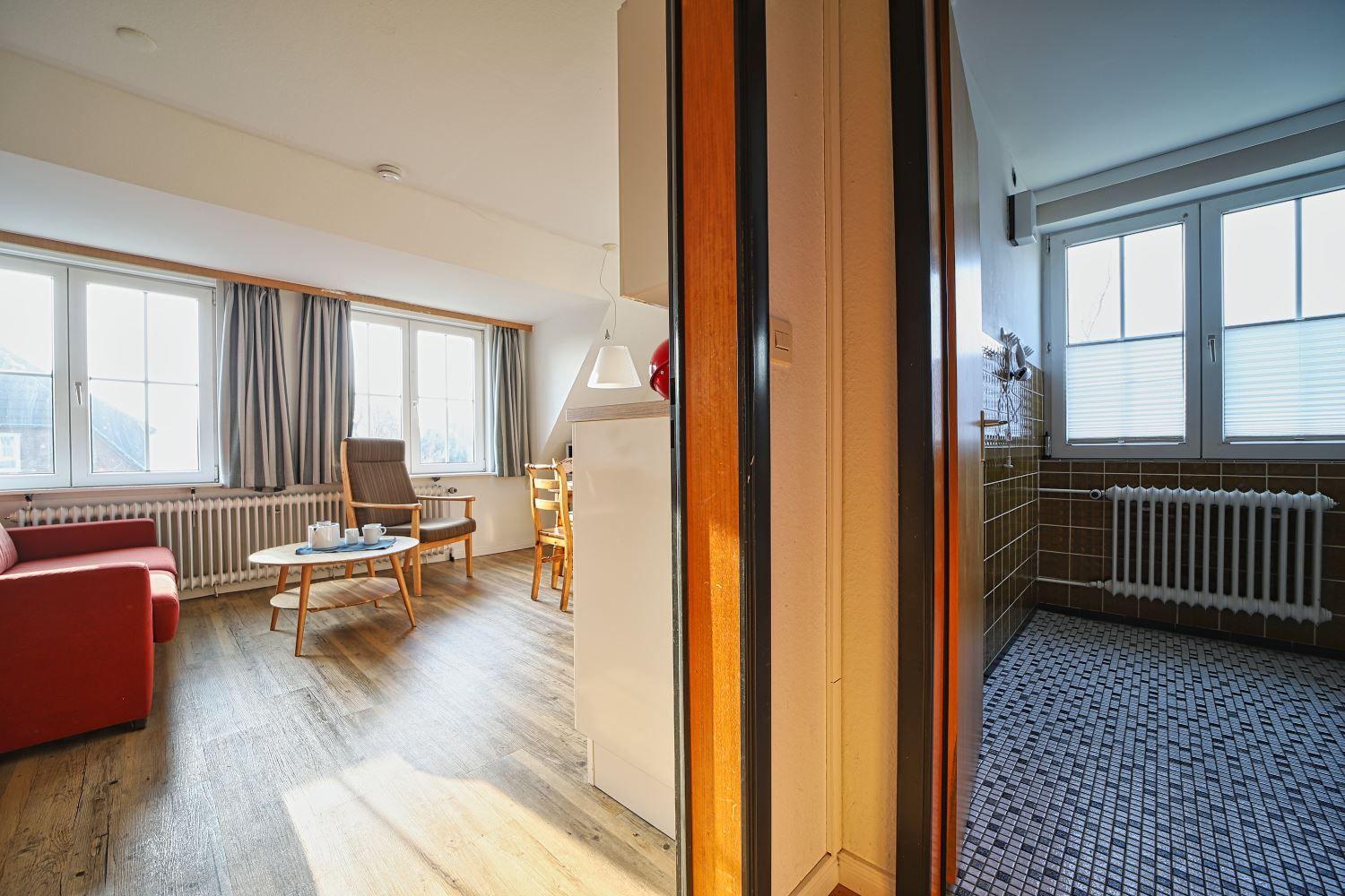 Wohnzimmer mit Sofa, Sessel und Fenstern. Nebenraum mit Fliesenboden und Fenster.