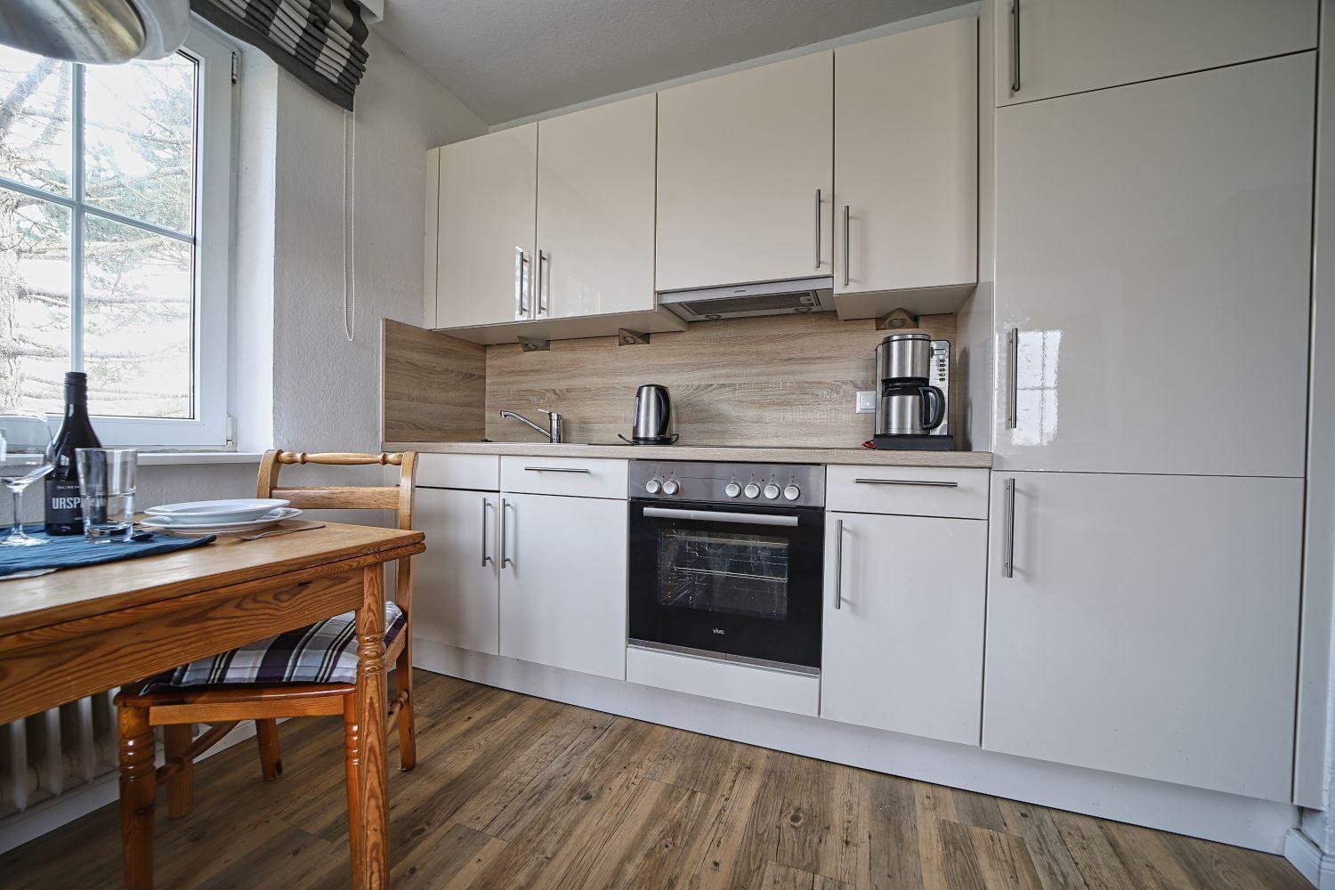 Küche mit Essplatz, weißen Schränken und Holzboden. Fenster mit Blick nach draußen.