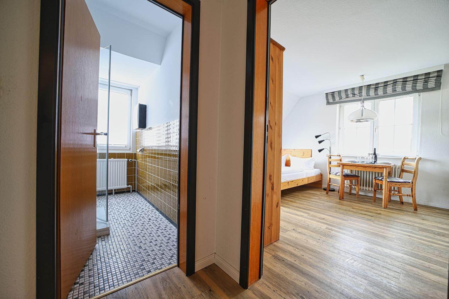 Zimmer mit Holzboden, Bett, Tisch und Stühlen. Nebenraum mit Dusche und Fliesen.