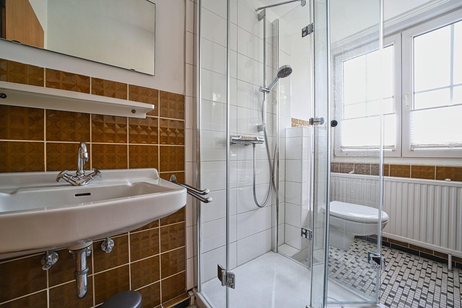 Bad mit Dusche, Waschbecken und Toilette. Fliesenwand und Fenster sichtbar.