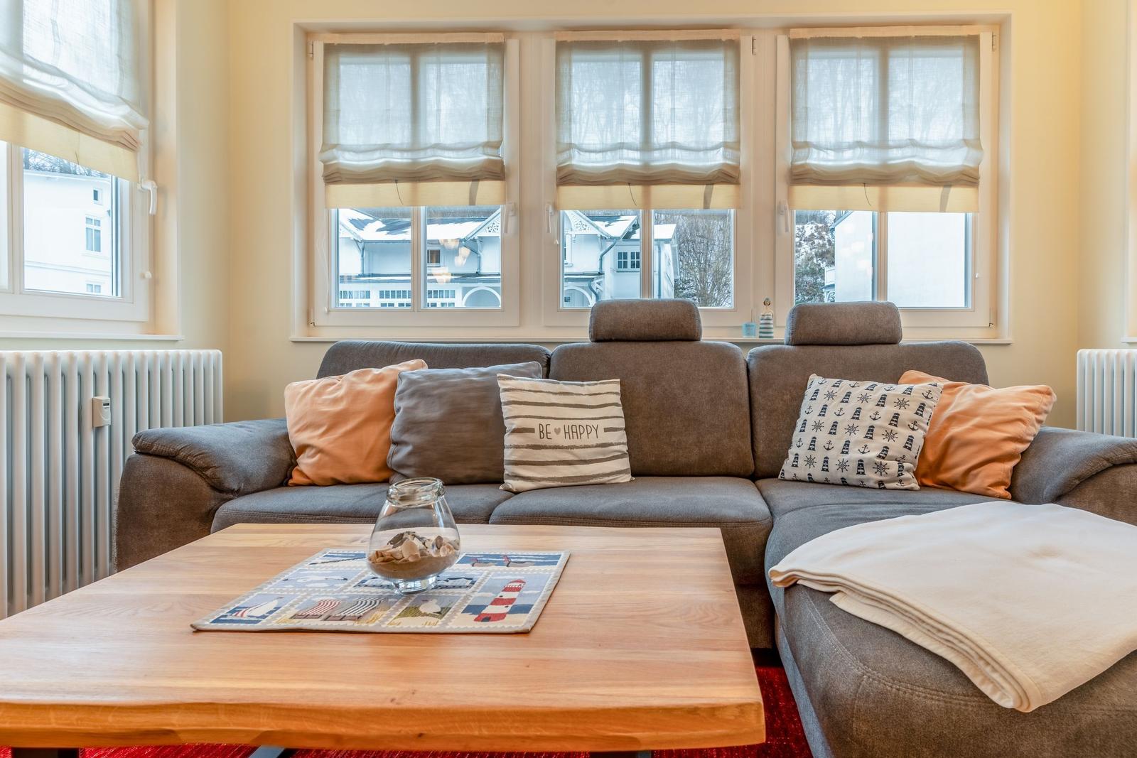 Ein Wohnzimmer mit grauem Sofa, Holztisch und Fenstern mit Rollo.