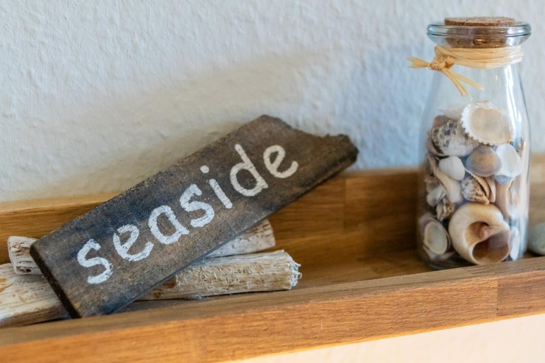 Holzschild mit Aufschrift seaside und Glasflasche mit Muscheln auf Holzregal.