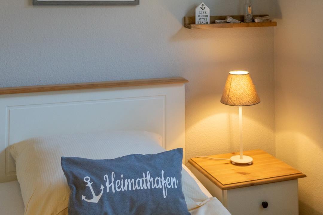 Schlafzimmer mit Bett, Nachttisch und Lampe. Kissen mit Aufschrift Heimathafen.